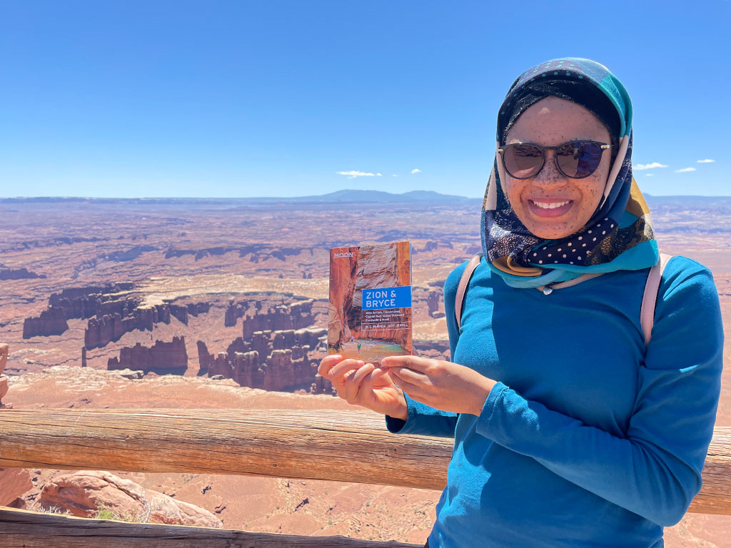 woman in a hijab and sunglasses posing with a book in front of the grand canyon